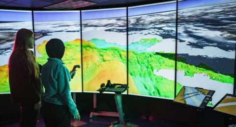 Two researchers stand before a wall-sized screen showing ocean mapping data